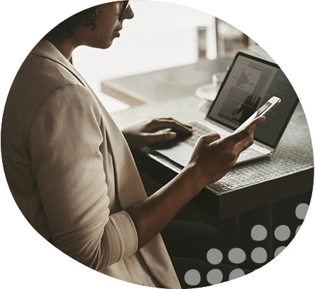 Photo of lady sat looking at mobile, while working on laptop at desk